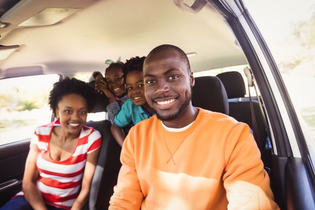 This image shows a joyful African American family posing together inside a car. The father is in the driver's seat, smiling at the camera, while the mother and children are in the back, also smiling. This photo is perfect for use in advertisements, blogs, or articles about family bonding, road trips, vacations, and joyful moments. It can also be used in promotional materials for car companies, travel agencies, or family-oriented products and services.