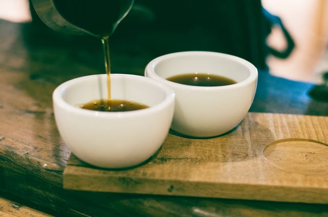 A close-up image showing coffee being poured into two white ceramic cups placed on a wooden trivet. Ideal for promoting cafes, coffee shops, barista training, morning routines, hot beverages, cozy environments, and relaxation moments.
