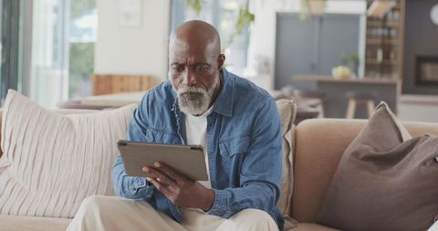 Senior african american man using tablet on sofa reading news and relaxing in modern home