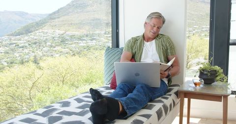 Mature man working remotely on laptop and taking notes while reclining by window with tea
