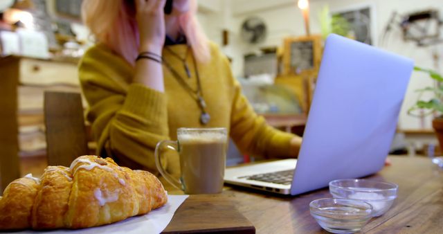 Young woman sitting in a cozy cafe, working on her laptop while sipping coffee. Next to her is a croissant on a napkin, adding to the relaxed ambiance. The setting suggests a freelance work environment, ideal for themes of remote work, digital nomad lifestyle, and casual workspaces.
