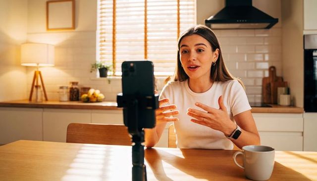 Content creator recording lifestyle vlog at kitchen table with smartphone tripod and mug