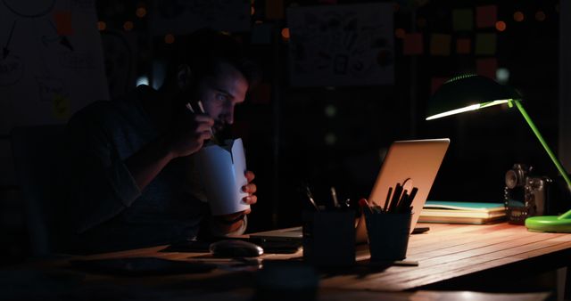 A man working late at night in an dimly lit office. He is sitting in front of a laptop, eating takeout food from a white container. Desk covered with stationery, pencils, documents, and a desk lamp casting soft light on the scene. Ideal for illustrating themes of late-night work, dedication, urban office life, work-life balance, and business environments.