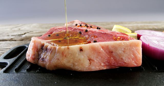 Raw steak sprinkled with black pepper and being drizzled with olive oil, ready for cooking. Slice of red onion and lemon in background. Ideal for use in food blogs, recipe websites, and gourmet cooking promotions.