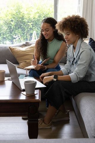 African american women collaborating on laptop at home, taking notes and planning together