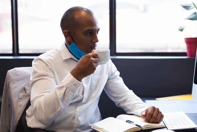 Biracial businessman wearing lowered face mask drinking coffee sitting at desk. health and hygiene in workplace during coronavirus covid 19 pandemic.