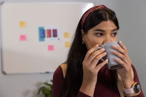 Asian mid-adult woman drinking coffee in modern office near whiteboard with sticky notes