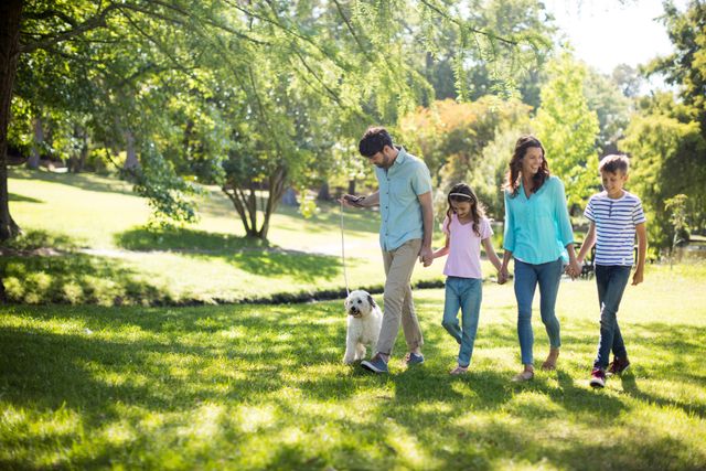 Family of four with dog walking in park on sunny day. Parents and children holding hands, enjoying nature and bonding. Ideal for use in advertisements, family-oriented content, lifestyle blogs, and promotional materials for outdoor activities.