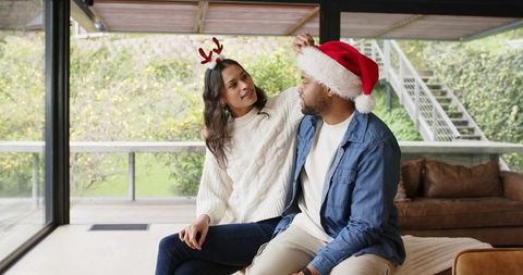 Diverse couple sitting on sofa adjusting santa hat while woman wears antler headband