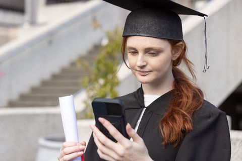 Redhead graduate in cap and gown checking smartphone while holding diploma on campus