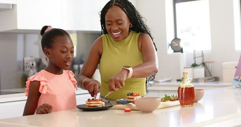 Smiling mother and daughter preparing stacked berry pancakes with syrup for breakfast