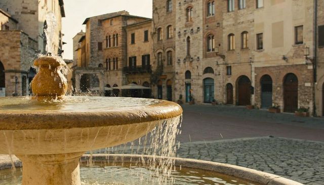 Sunlit tuscan town fountain spilling water over stone basin in historic cobblestone piazza