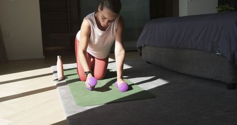 Woman kneeling on mat gripping dumbbells in sunlit bedroom during home workout