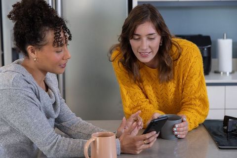 Multiracial friends leaning and browsing smartphone at bright modern kitchen counter