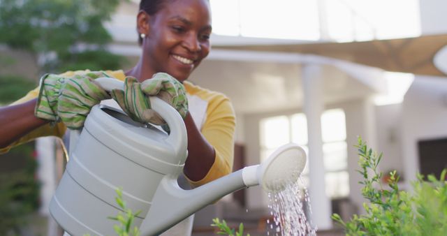 Happy african amercian woman wearing gloves watering flowers in garden. staying at home in self isolation during quarantine lockdown.