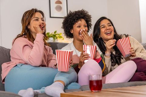 Laughing diverse friends watching movie, sharing popcorn and drinks, cozy home night