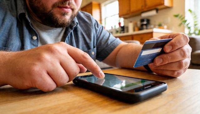 Bearded man making mobile payment with smartphone and credit card at kitchen table