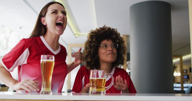 Two women in sports jerseys are in a bar celebrating and cheering while watching a sports game. They are drinking beer and displaying emotions of excitement and joy. This image can be used for promotions of sports events, advertisements for bars or pubs, or campaigns targeting sports fans.