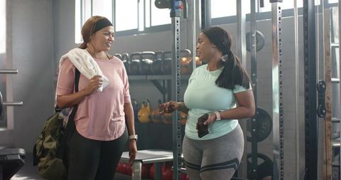 Plus size friends chatting after workout in bright gym with kettlebells and squat rack
