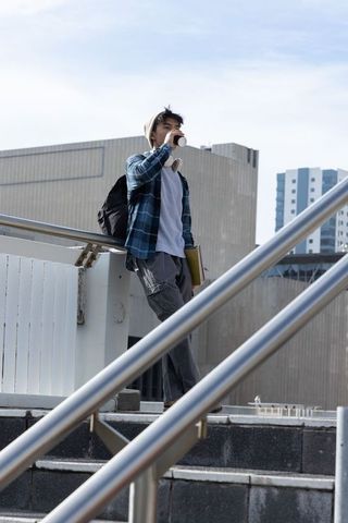 Asian student sipping coffee on city overpass with backpack, headphones, notebooks