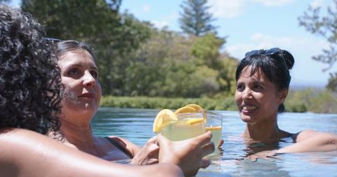 Three women clinking lemon cocktails in infinity pool enjoying sunny outdoor relaxation