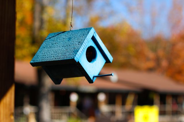 Blue wooden birdhouse hanging outdoors with blurred background of autumn trees and rustic houses. Ideal for themes such as bird watching, nature, outdoor decor, and seasonal change. Perfect for editorial use in articles about gardening or bird conservation, or as simple artwork for home decor.