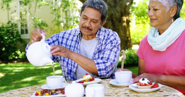A middle-aged Latino man pours tea while a middle-aged woman watches, both enjoying an outdoor tea time, with copy space. They are sharing a moment of leisure in a garden setting, reflecting a relaxed and pleasant lifestyle.