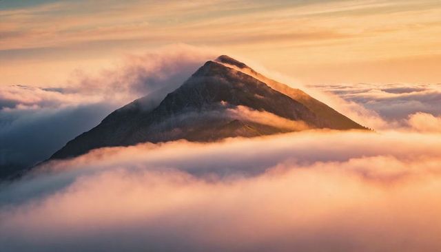 Triangular mountain peak piercing sea of clouds at golden hour, alpenglow lighting ridge