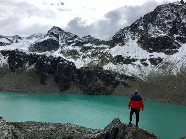 Hiker standing on rocky terrain gazing at tranquil turquoise mountain lake surrounded by snow-capped peaks. Ideal for use in promotional materials for adventure travel, hiking tours, nature exploration brochures, and outdoor lifestyle content.