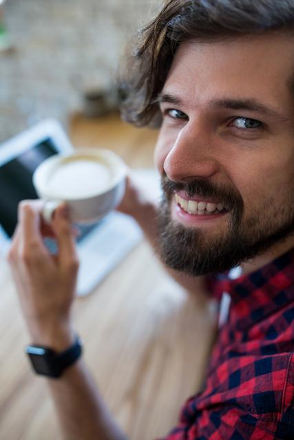 Young man with beard smiling while holding a cup of coffee in a cafe. He is sitting at a wooden table with a laptop in front of him, suggesting a relaxed and casual atmosphere. Ideal for use in lifestyle blogs, remote work articles, coffee shop promotions, and advertisements focusing on leisure and happiness.