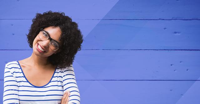 Young woman with curly hair and glasses standing against a blue wooden background, smiling confidently. Ideal for use in lifestyle blogs, fashion websites, advertisements, and social media posts promoting positivity and confidence.