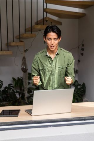 Asian man celebrating remote work win at modern home workspace with laptop and plants