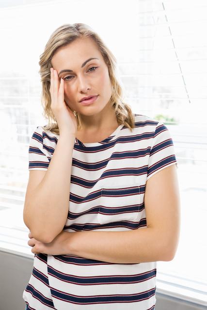 Young woman standing indoors, appearing stressed and deep in thought. She is wearing a striped shirt and leaning against a window with natural light streaming in. This image can be used to depict themes of mental health, emotional struggles, stress, and contemplation. Suitable for articles, blogs, and campaigns related to mental health awareness, personal stories, and emotional well-being.