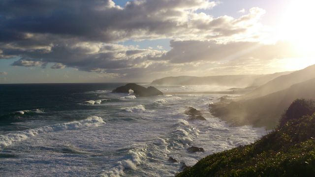Dramatic coastal scene with sunlit clouds and waves crashing into rocky shoreline. Suitable for themes of nature, travel, weather, and serenity. Ideal for backgrounds, website headers, travel blogs, brochures, and educational materials on coastal environments.