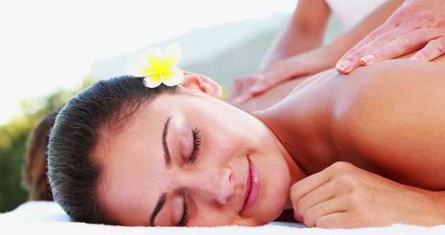 Image depicts a woman with a tranquil expression relaxing outdoors while receiving a back massage. A flower is tucked behind her ear, enhancing the serene ambiance. This visuals is ideal for advertisements for spa services, wellness and self-care blogs, or promotional materials for health resorts.