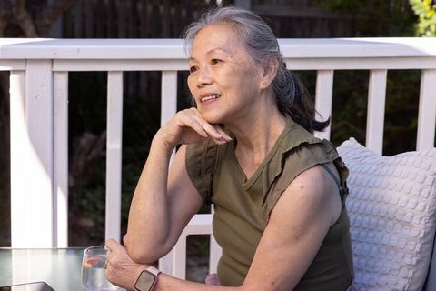 Senior asian woman sitting on sunlit deck resting chin wearing smartwatch and olive top