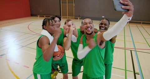Diverse male basketball team celebrating and taking group selfie on indoor court after win