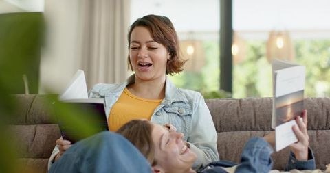 Friends relaxing on sofa reading together, laughing and sharing stories at home