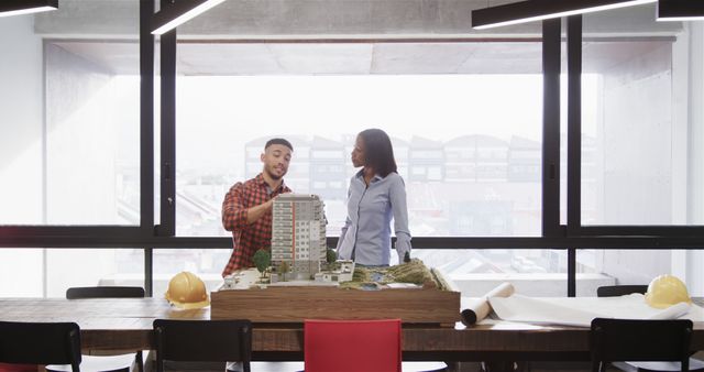 Two architects are discussing a building model in a modern office, with hard hats and blueprints on the table. This can be used for themes like teamwork, architecture, construction planning, engineering projects, and collaborative work environments.