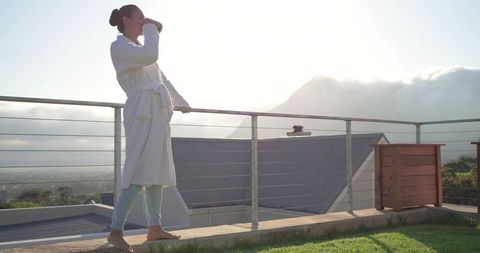 Asian woman sipping morning coffee on rooftop terrace at sunrise wearing white bathrobe