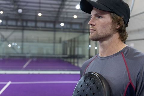 Athlete standing beside purple padel court holding paddle wearing cap focused gaze