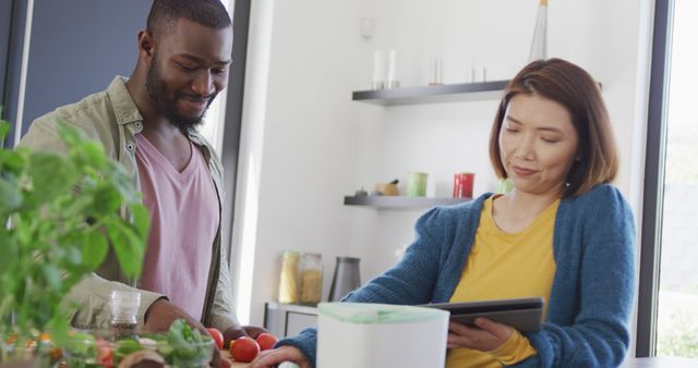 Image of happy diverse couple preparing food, using tablet and composting vegetables in kitchen. Domestic life, communication, togetherness, health, happiness and inclusivity concept.