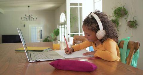 African american girl studying at dining table wearing headphones writing with laptop