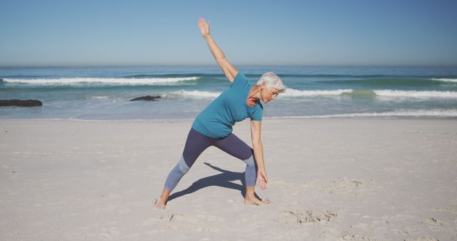 Senior woman engaging in yoga exercises on a sandy beach under clear blue sky. Ideal for use in promoting healthy lifestyles, fitness programs for the elderly, wellness and mental health initiatives, and advertisements targeting senior activity programs. Perfect for articles on staying active in later life, maintaining health and fitness, and enjoying outdoor environments.