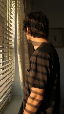 Vertical video of man by window with blinds casting sunlit shadows, contemplative mood