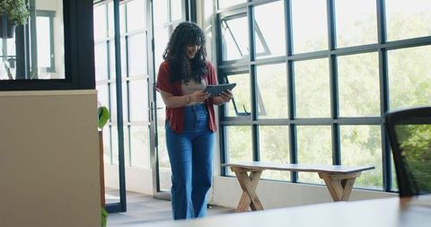 African american woman walking and using tablet in sunlit modern office with grid windows