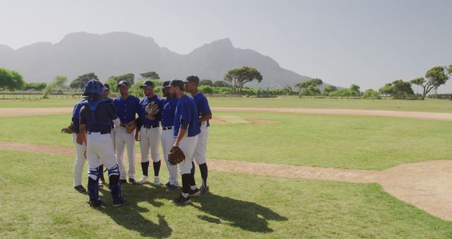 Group of baseball players in blue uniforms huddling on an open field with mountains in the background under a clear sky. Ideal for use in sports articles, teamwork motivation, outdoor activities promotions, athletic events advertising, and community sports programs.