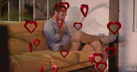 Red-haired man sitting on mustard sofa crossing legs and gazing amid floating red hearts