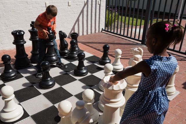 Children engaging in a game of giant chess outdoors on a sunny day. Ideal for illustrating concepts of learning through play, teamwork, and outdoor activities in educational settings. Can be used in educational materials, school brochures, and articles about children's games and outdoor activities.