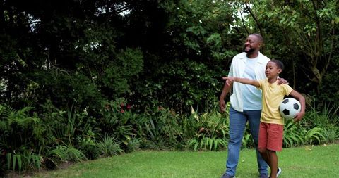 Dad guiding son during backyard soccer practice holding ball and pointing toward goal